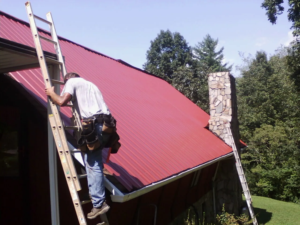 Roofer on a ladder beside a red metal roof and stone chimney — Soffit & Fascia Repair by Roofing Brothers in Marthasville